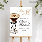 A personalized third birthday party sign styled as a cocktail menu card, displayed on a wooden easel. The white card features a watercolor martini glass with coffee beans and a black ribbon, alongside elegant text reading 'a tini bit older' and 'WELCOME TO Olivia's Thirtieth BIRTHDAY PARTY EST.
