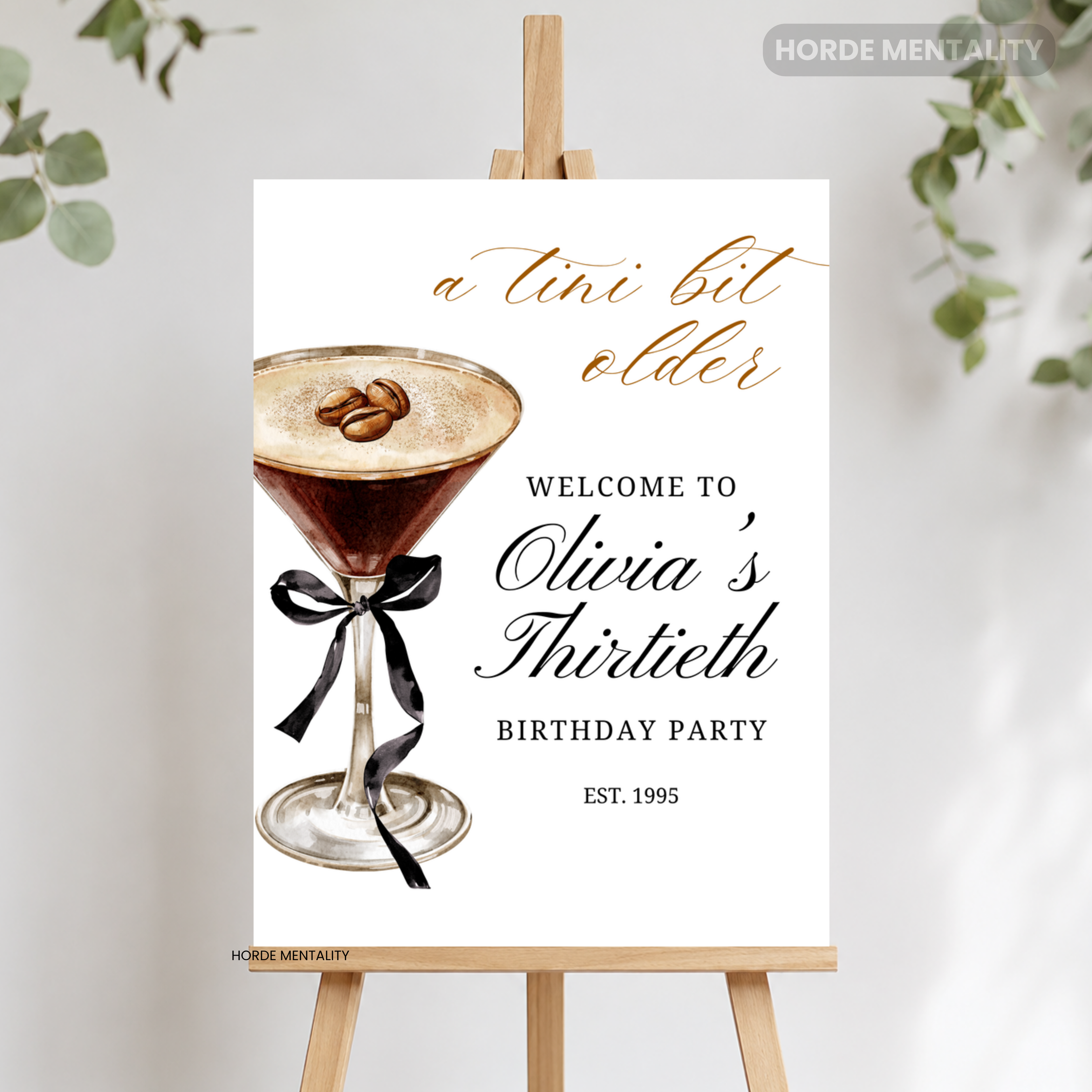 A personalized third birthday party sign styled as a cocktail menu card, displayed on a wooden easel. The white card features a watercolor martini glass with coffee beans and a black ribbon, alongside elegant text reading 'a tini bit older' and 'WELCOME TO Olivia's Thirtieth BIRTHDAY PARTY EST.