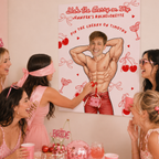 A group of women at a bachelorette party are gathered around a large personalized bachelorette canvas print featuring a muscular cartoon man and red text. The room is decorated in pink tones with party cups, cherries, and a pink mirror on the table.