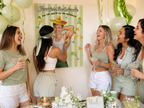 A group of five women laughing and celebrating at a bachelorette party, gathered around a large personalized printed poster that reads 'Jennifer's Bachelorette' and 'Pin the nacho on muchacho.' The scene includes balloons, drinks, flowers, and party decor, making it a clear fun girls bach party