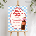 A personalized baby shower sign on a wooden easel, featuring a blue and white checkered border, a red scalloped frame, the text 'She's Ready to Pop! Welcome to Amanda's Baby Shower,' a printed bottle illustration with a pink bow